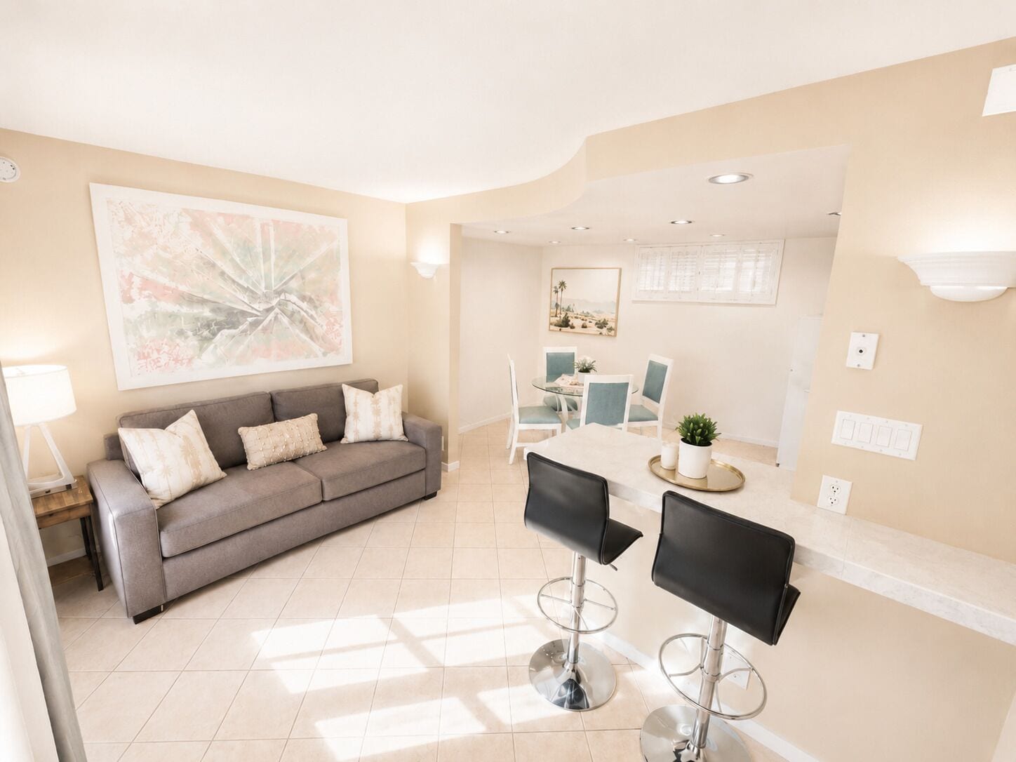 A modern living room with a grey sofa, a dining area with a table and chairs, and a kitchen area with a counter and stools.