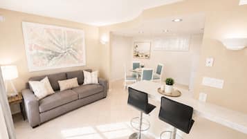 A modern living room with a grey sofa, a dining area with a table and chairs, and a kitchen area with a counter and stools.