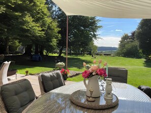 Une terrasse extérieure avec des meubles en osier, une table agrémentée d’une bougie et de fleurs, et une vue sur un espace verdoyant avec des arbres.