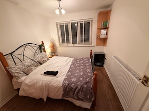 Un dormitorio con cama, lámpara de noche, ventana, radiador y estantería con libros.