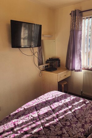 Un dormitorio con cama, escritorio con computadora, televisor montado en la pared y ventana con cortinas.