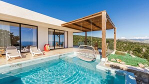 Un área de piscina exterior moderna con agua azul cristalina, una pérgola de madera y vistas al paisaje.