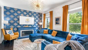 Un salón con un sofá azul, un sillón amarillo, una chimenea y un ventanal con cortinas.