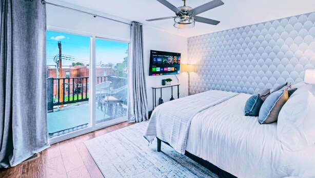 A bedroom with a bed, a ceiling fan, a TV, and a balcony with a view of buildings. - Hot Tub, Games, Gated Parking near SLU & Downtown (St. Louis)