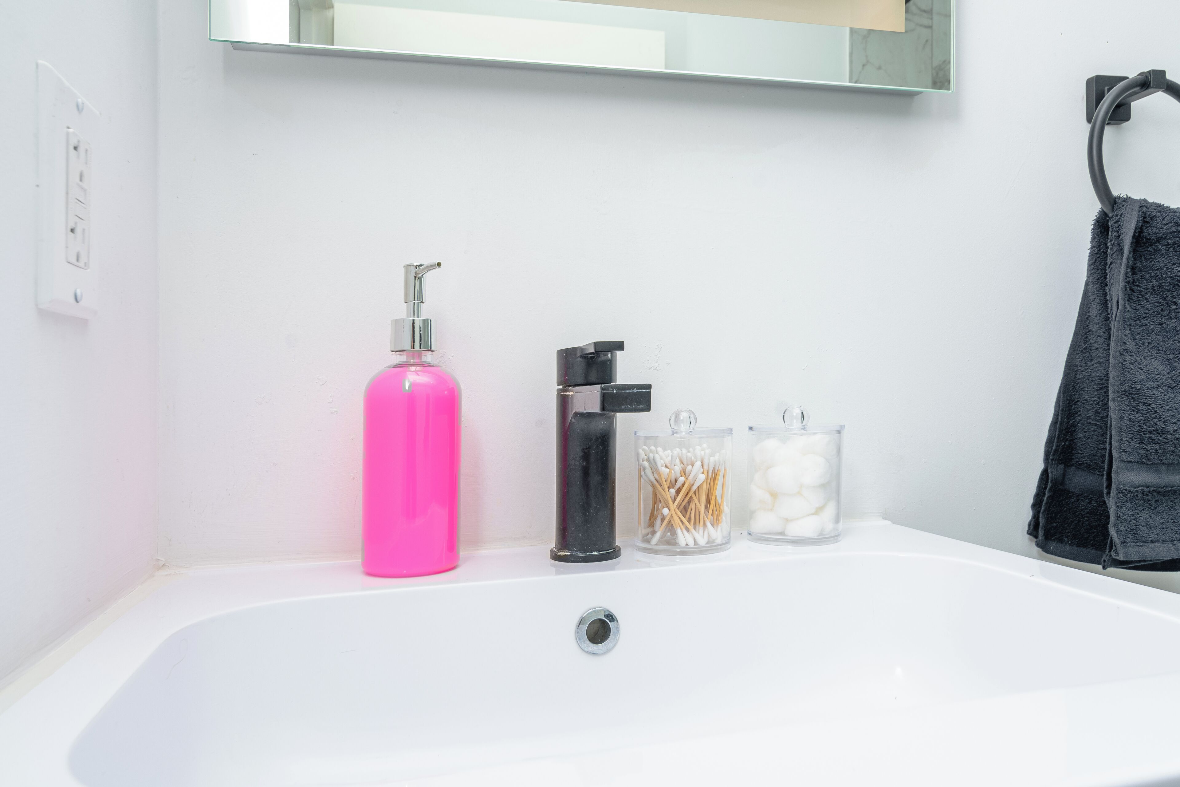 A bathroom with a white sink, a pink bottle, a black soap dispenser, and two glass containers with cotton balls and cotton swabs.