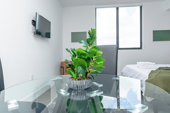 A modern room with a glass table, a potted plant, a bed with a white bedspread, and a TV mounted on the wall.