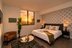 Une chambre d’hôtel avec un lit, une chaise, une table et une grande fenêtre donnant sur un magnifique coucher de soleil.