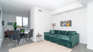 A modern living room with a green velvet sofa, a dining area with a table and chairs, and a large window providing natural light. - Central 1BR in Miami Near Wynwood and Marlins Park (Miami)