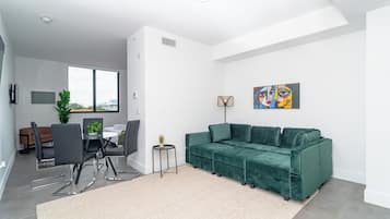 Ein modernes Wohnzimmer mit einem grünen Samtsofa, einem Essbereich mit Tisch und Stühlen sowie einem großen Fenster, das für natürliches Licht sorgt.