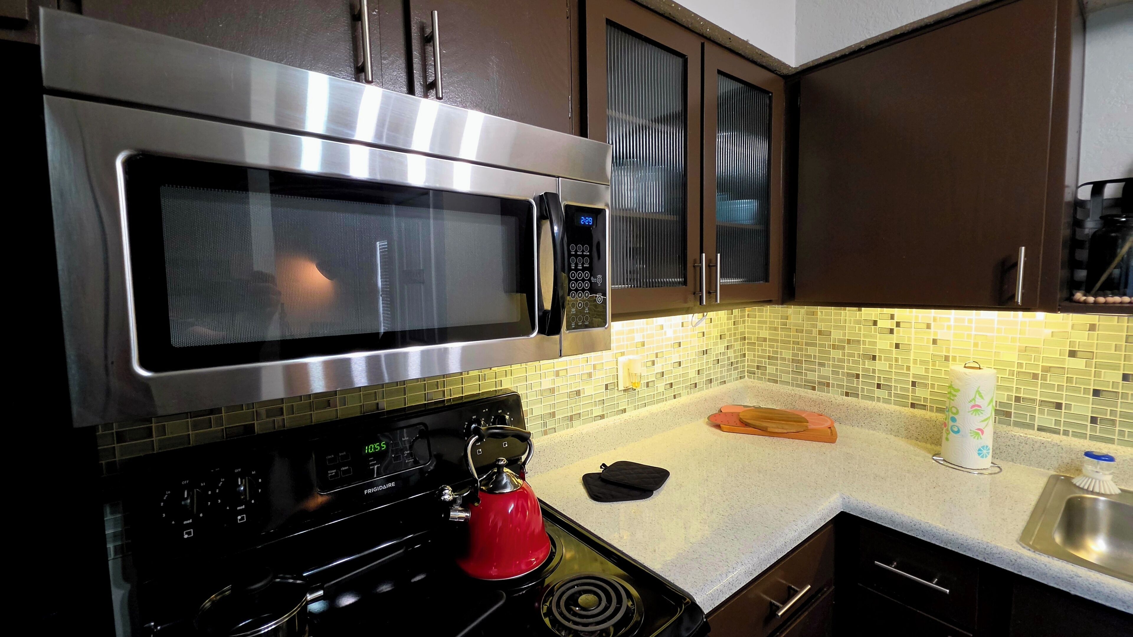 Una cocina con microondas de acero inoxidable, un hervidor rojo, un reloj digital y un salpicadero de azulejos.