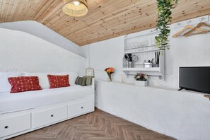 A cozy room with a white sofa, a TV, a wooden ceiling, and a shelf with kitchen utensils.