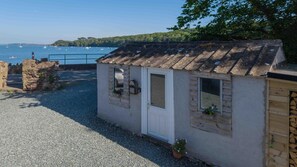 A small, single-story building with a tiled roof, a door, and windows, situated on a gravel path with a view of the sea and boats in the background.