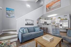 Ein modernes Wohnzimmer mit einem blauen Sofa, einem Holztisch und einer Küchenzeile mit weißen Schränken und Edelstahlgeräten.