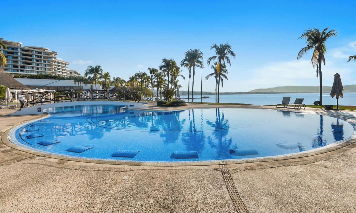 Ein kreisförmiger Infinity-Pool mit klarem, blauem Wasser, umgeben von Palmen und Liegestühlen, mit Blick auf das Meer und einem Hotel im Hintergrund.