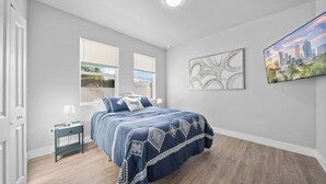 Un dormitorio con cama, dos ventanas, una mesita de noche, lámpara, un cuadro abstracto enmarcado y una pintura de paisaje urbano en la pared.