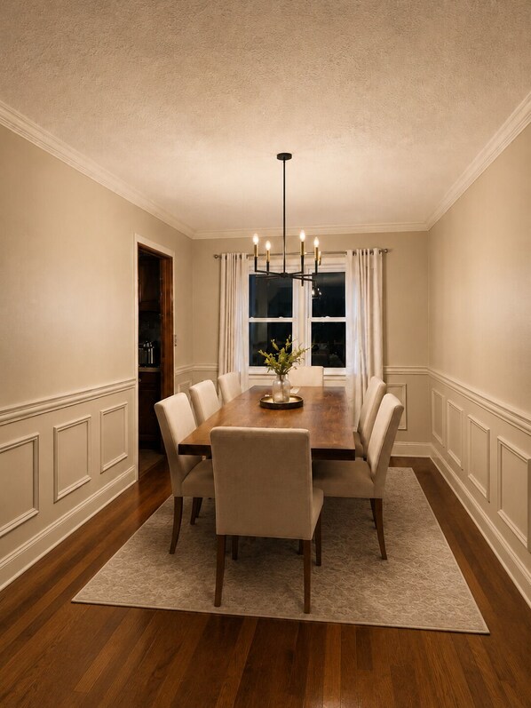 A dining room with a wooden table, six chairs, a chandelier, and a window with curtains.