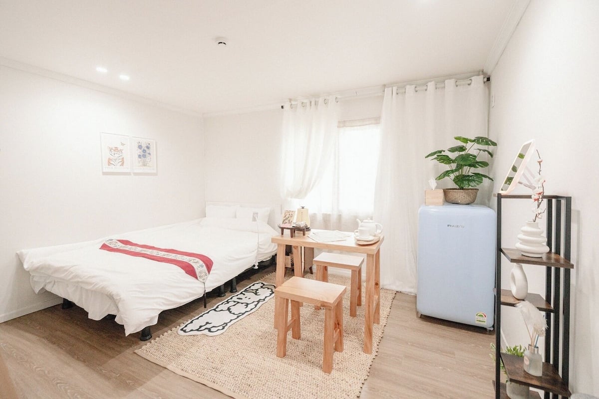 Quarto pequeno e limpo com cama, escrivaninha, cadeira, frigorífico e planta em vaso.