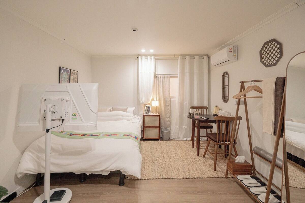 Quarto de hotel com cama, mesa de cabeceira, cadeira, escrivaninha e uma pequena mesa com luminária.