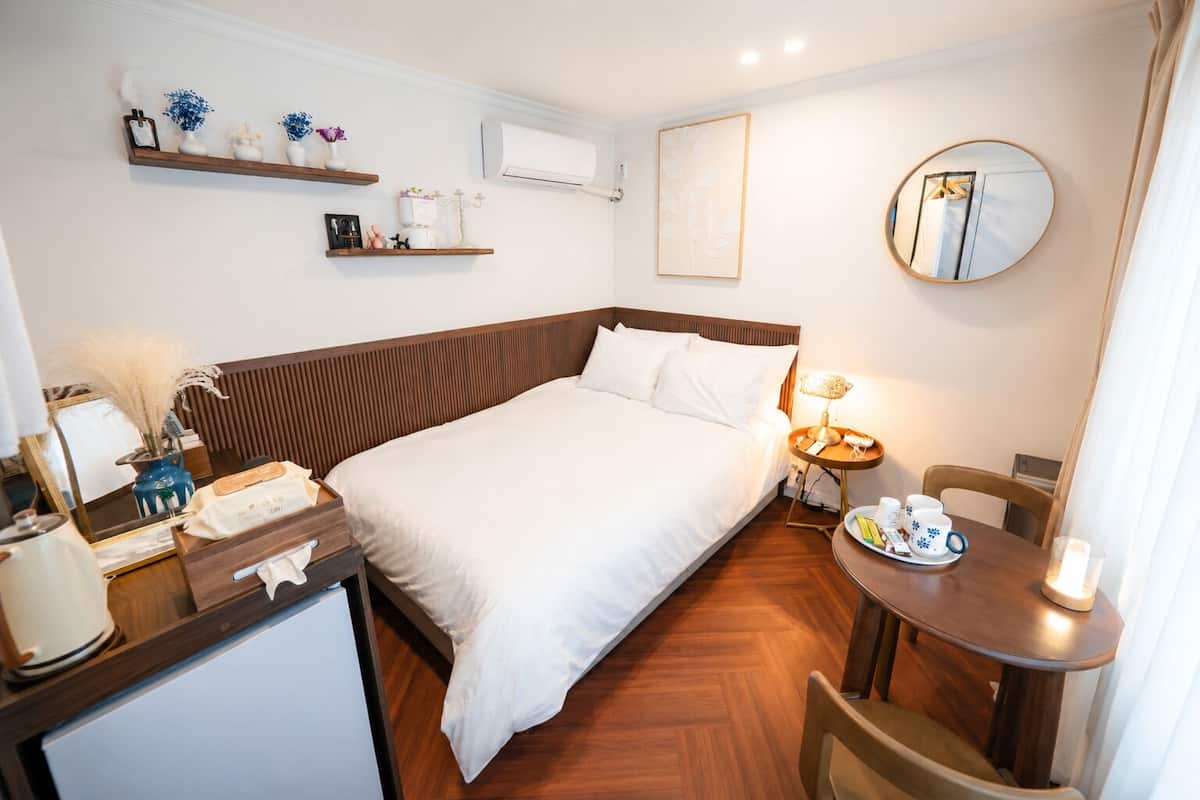 Quarto de hotel com cama, mesa de cabeceira, escrivaninha, cadeira e pequena mesa com xícaras e uma vela.