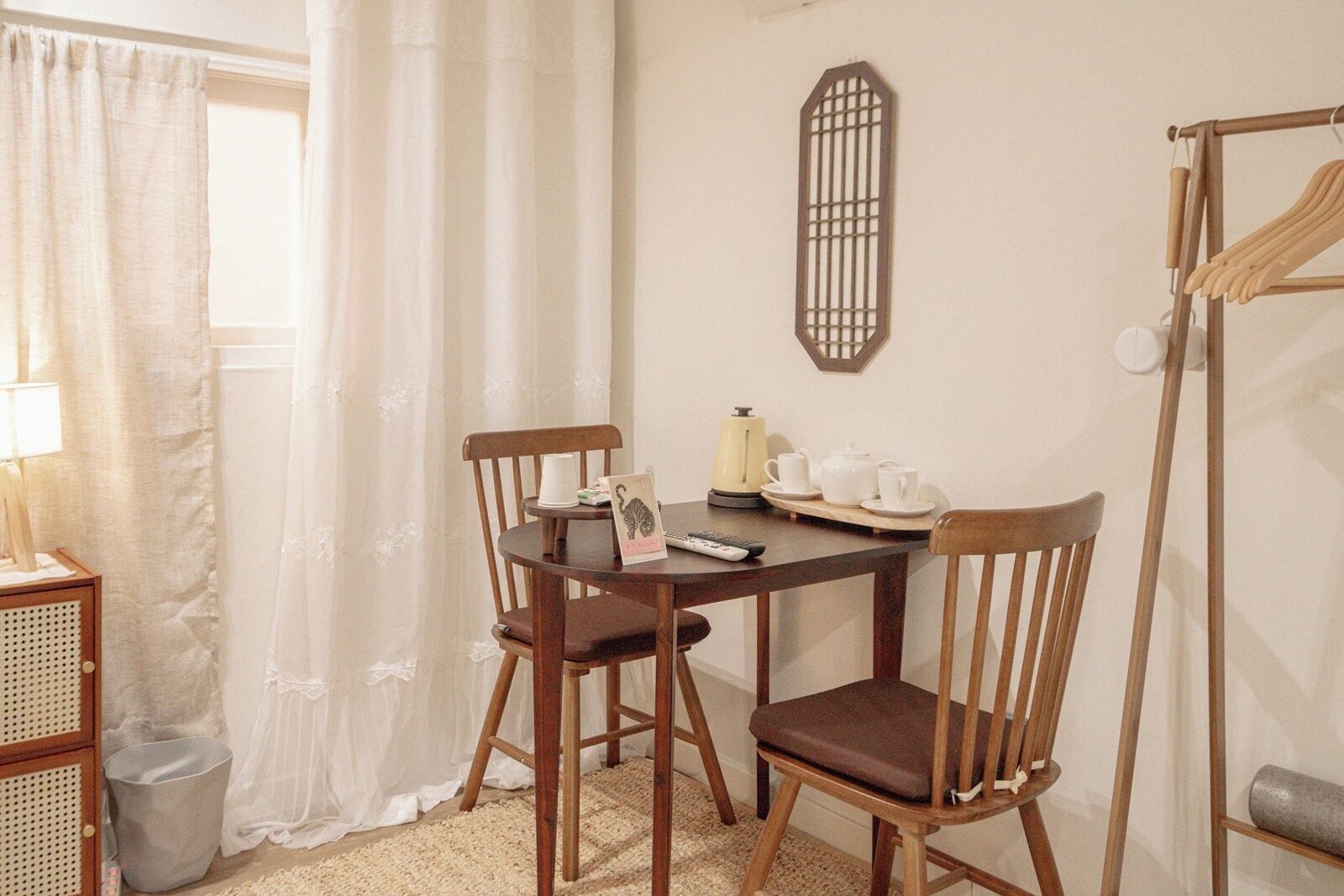 A room with a round wooden table, two wooden chairs, a small cabinet, a radiator, a wall-mounted mirror, and a window with sheer curtains.