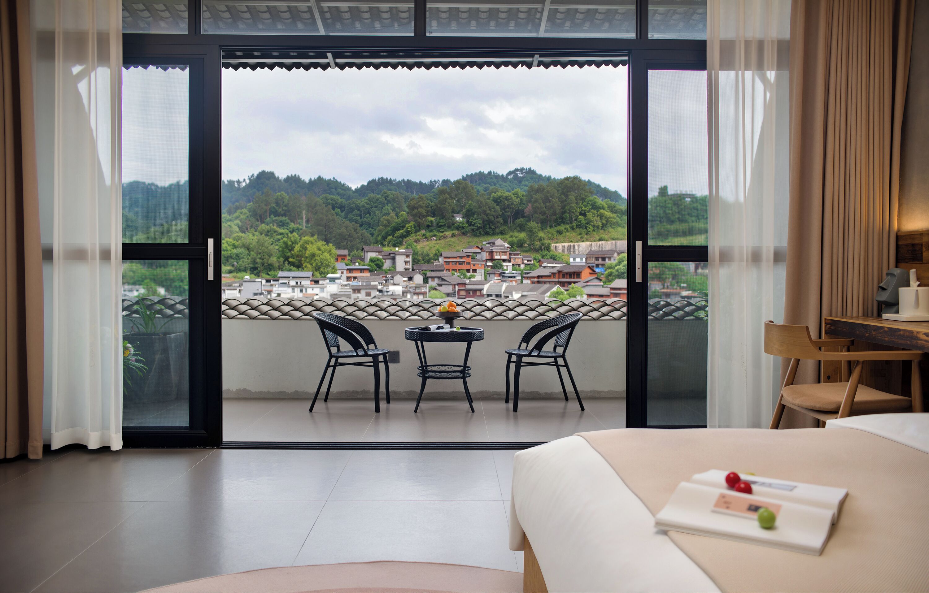 A hotel room with a balcony, a bed, a table with chairs, and a view of a village.