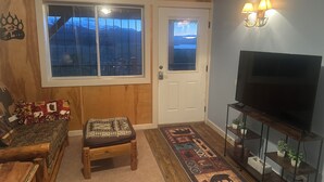 Un accogliente soggiorno con divano, una piccola panca in legno, un televisore su un mobile, una porta con vista e un lampadario a parete.