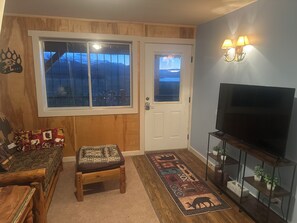 Ett mysigt vardagsrum med en soffa, en liten träbänk, en tv på ett tv-bord, en dörr med utsikt och en väggmonterad lampa.