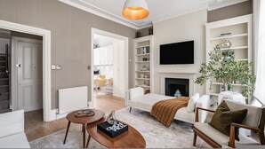 A living room with a fireplace, a TV mounted on the wall, and a dining area in the background. - 5-bed Chelsea Home Next To Kings Road (London)