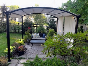 Un área de patio techada con un tapete estampado, un sofá con cojines y plantas en macetas.