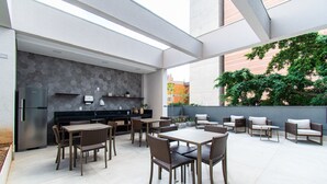 A modern outdoor dining area with a kitchen, seating, and a view of the city. - Pause Living - Nex One Paraíso (São Paulo)