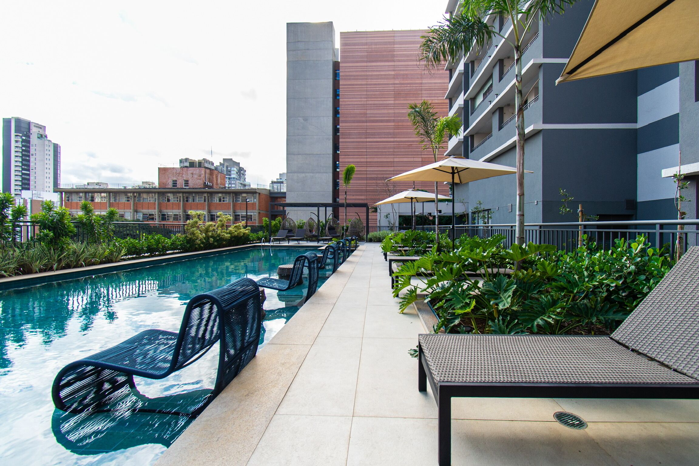 A pool area with lounge chairs and umbrellas, surrounded by greenery and modern buildings.