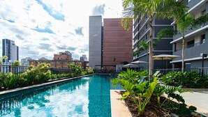 A swimming pool with a clear blue water, surrounded by greenery and a modern building in the background. - Pause Living - Nex One Paraíso (São Paulo)