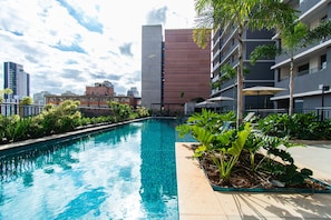 A swimming pool with a clear blue water, surrounded by greenery and a modern building in the background. - Pause Living - Nex One Paraíso (São Paulo)