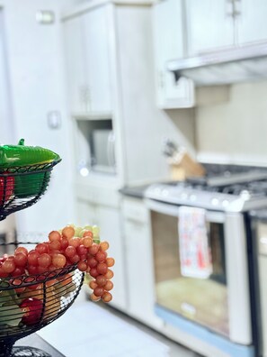 Une cuisine avec des meubles blancs, un fourneau et un panier de fruits contenant des raisins.