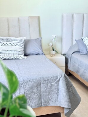 Una habitació amb dos llits, cadascun amb un llençol i coixins grisos, una taula de nit amb un ram de flors, i una petita taula auxiliar.