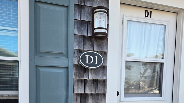 Une maison avec une porte grise portant l’inscription « D1 », une fenêtre avec un store blanc et une façade en bois.