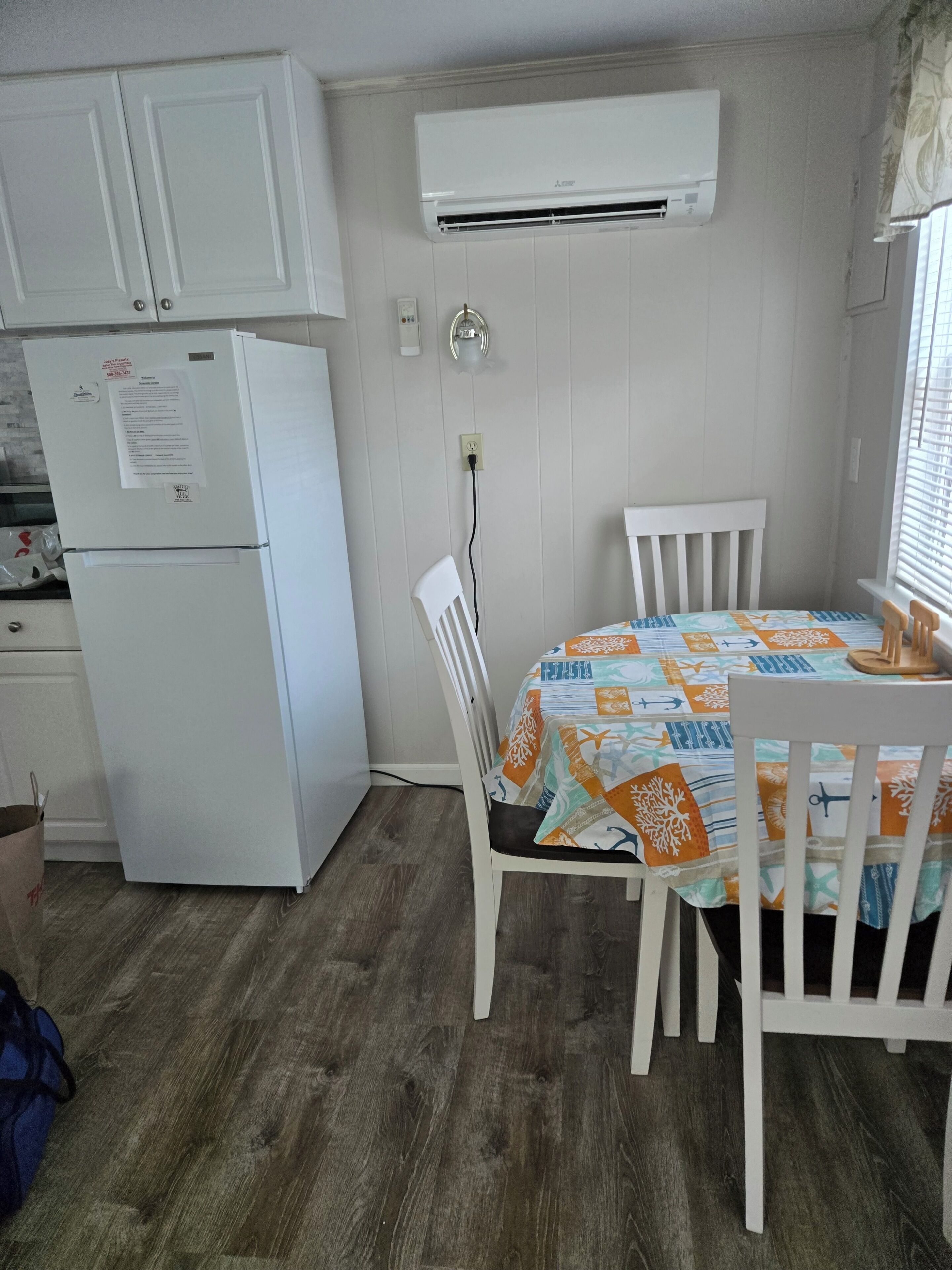 Una cocina compacta con un refrigerador blanco, un aire acondicionado instalado en la pared y un comedor con una mesa cubierta con un mantel estampado.