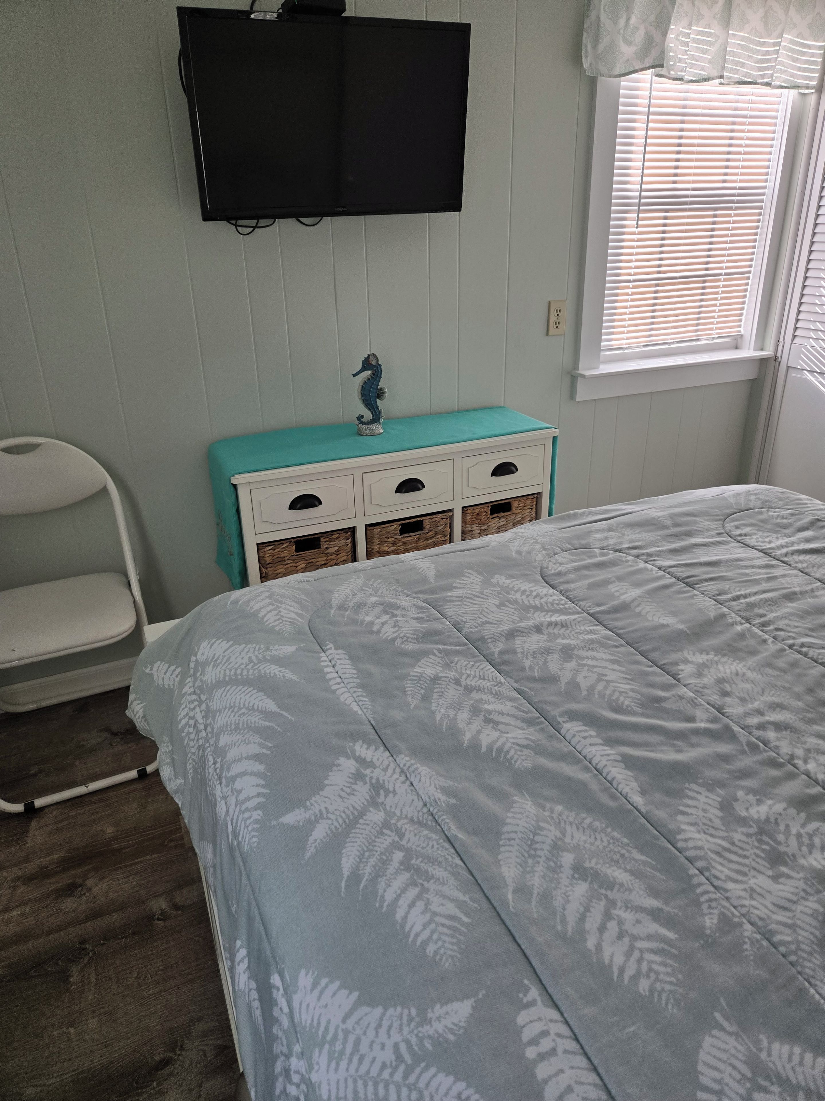 A bedroom with a bed, a dresser with drawers, a chair, a television mounted on the wall, and a window with blinds.