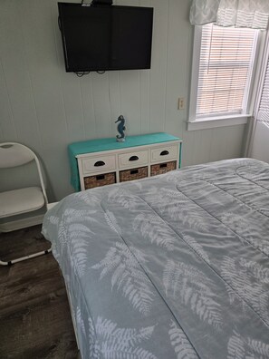 Un dormitorio con cama, cómoda con cajones, silla, televisor montado en la pared y ventana con persianas.