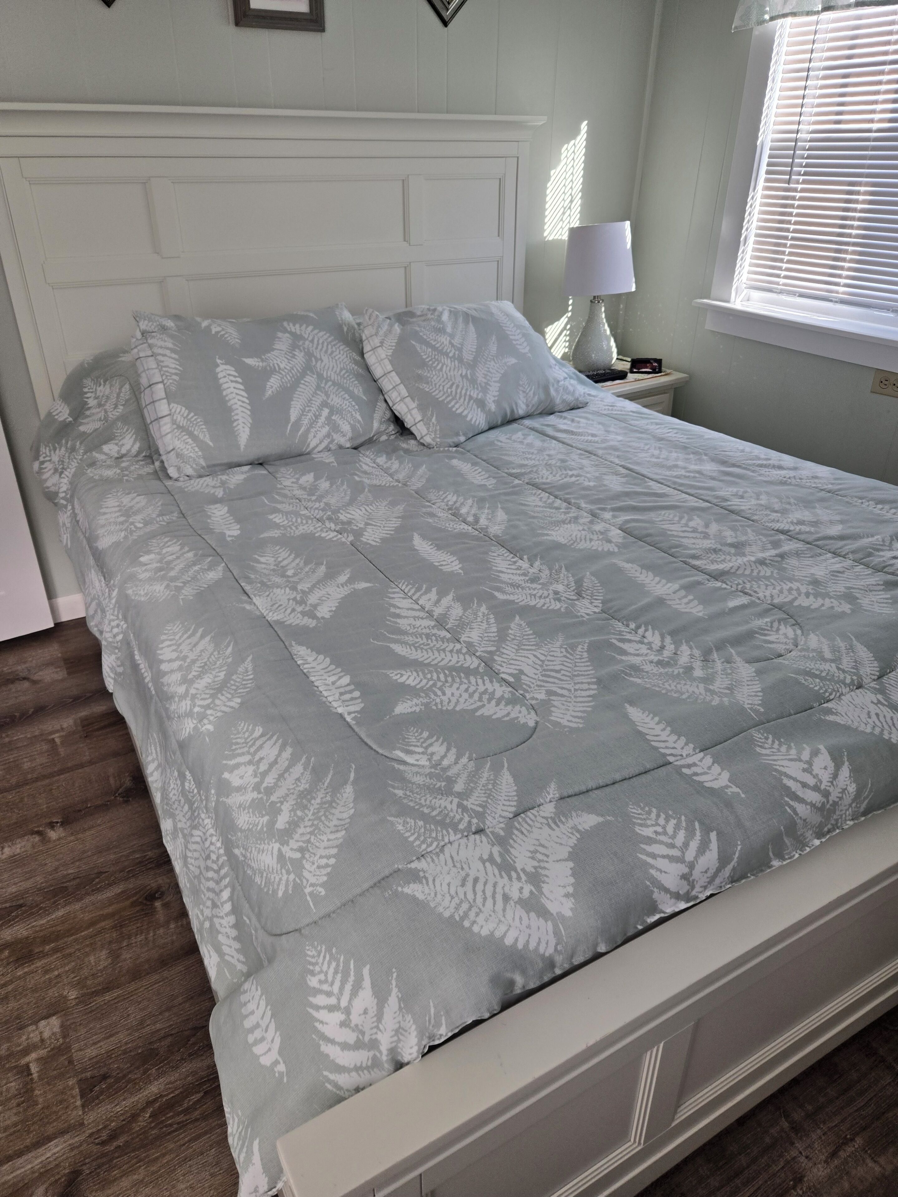 Una cama con colcha gris, una mesita de noche con lámpara y una ventana con persianas.
