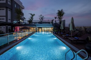 A rooftop pool with a bar area, lounge chairs, and palm trees.