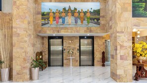A lobby with a stone wall, a painting of a temple, two elevators, and a small table with flowers. - Amana Hotel Phnom Penh (Phnom Penh)