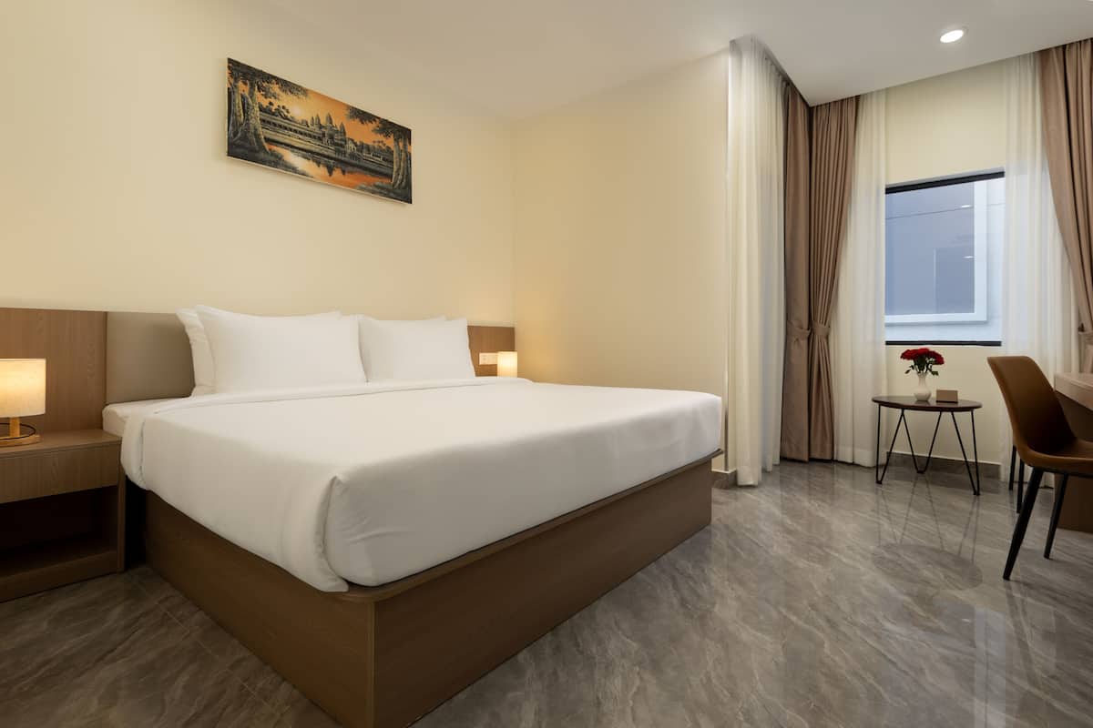 A hotel room with a large bed, a bedside table with a lamp, a small table with a vase, and a chair.