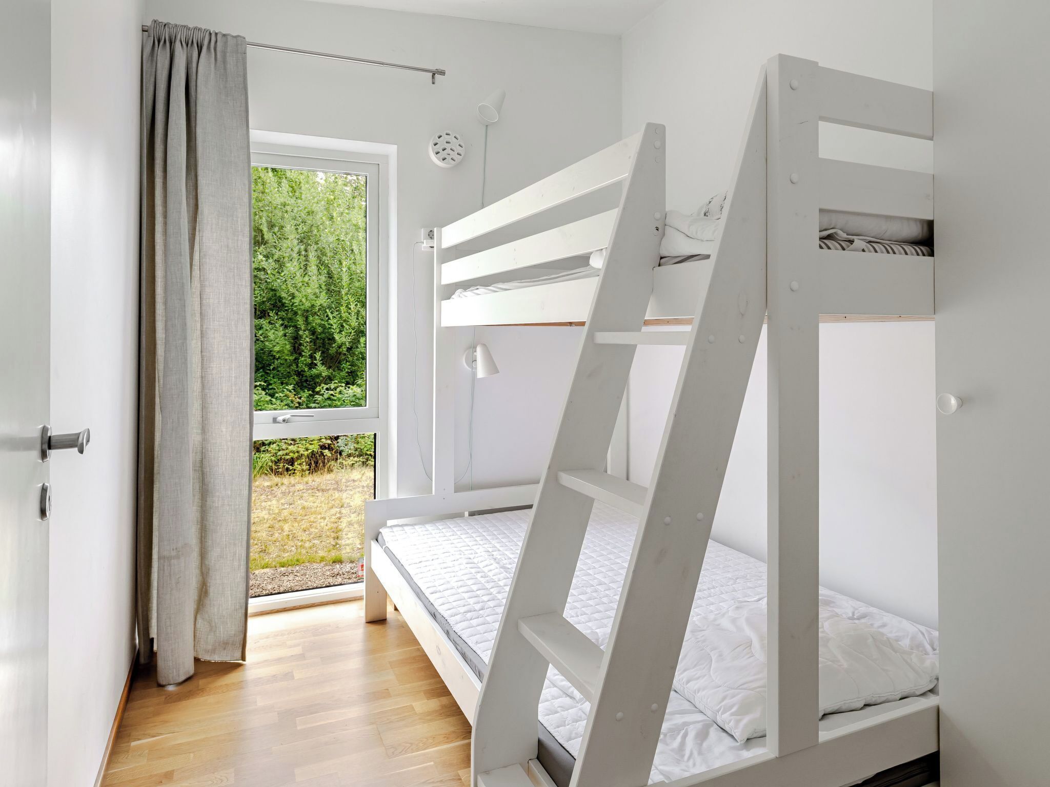A bedroom with bunk beds, a window with curtains, a white door, and a view of greenery outside.