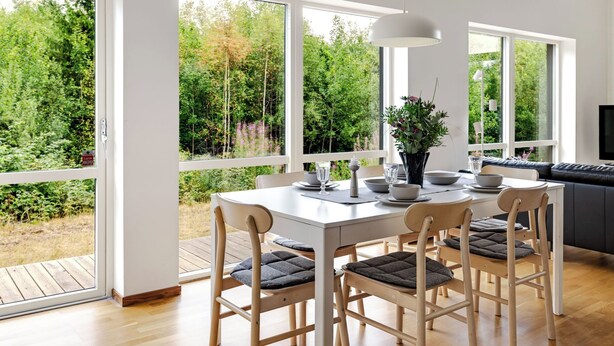 A modern dining area with a white table set for six, surrounded by large windows offering a view of a lush garden. - Paradiset 2 by Interhome (Holmsjö)
