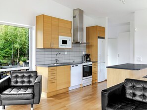 Eine moderne Küche mit Holzschränken, einer eingebauten Mikrowelle, einer Edelstahl-Dunstabzugshaube, einem Geschirrspüler und einem Kühlschrank. Zwei schwarze Ledersessel und ein Esstisch bieten Blick auf Grünfläche außerhalb.