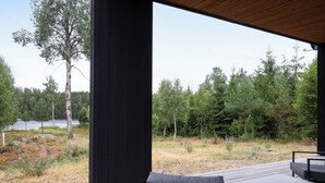 A deck with a black metal frame and a single grey cushioned chair, overlooking a forested area with a lake in the distance. - Paradiset 2 by Interhome (Holmsjö)