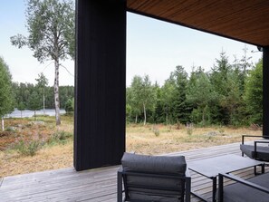 A deck with a black metal frame and a single grey cushioned chair, overlooking a forested area with a lake in the distance. - Paradiset 2 by Interhome (Holmsjö)