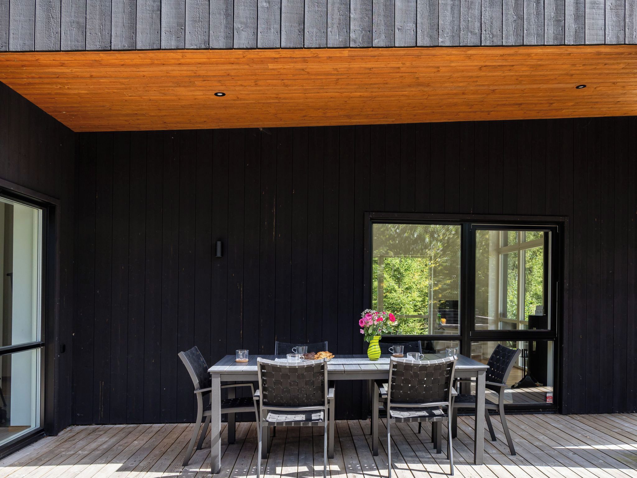 A modern outdoor dining area with a wooden deck, a table with chairs, and a large window offering a view of greenery.