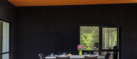 A modern outdoor dining area with a wooden deck, a table with chairs, and a large window offering a view of greenery.